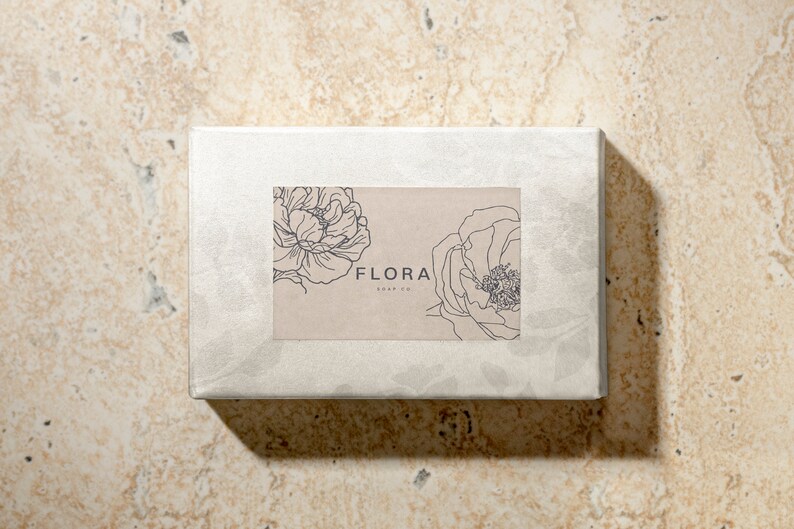 Puede incluir: Una caja blanca con una etiqueta marr&oacute;n que presenta un dibujo de l&iacute;nea de dos flores y el texto "FLORA SOAP CO."