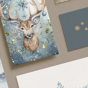 May include: A selection of Christmas greeting cards. One card displays a watercolour illustration of a deer head with antlers, snowflakes, and pear accents. Another card is blue with gold snowflakes and the text "A Happy Hol". A third card has "Merry Christmas" text.