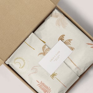 May include: A cardboard box with a white tissue paper lining. The tissue paper has a pattern of palm trees, leaves, and moons. A white business card with the text "Coco & Sand The Sweetest & Natural Beauty Balm" is on top of the tissue paper.