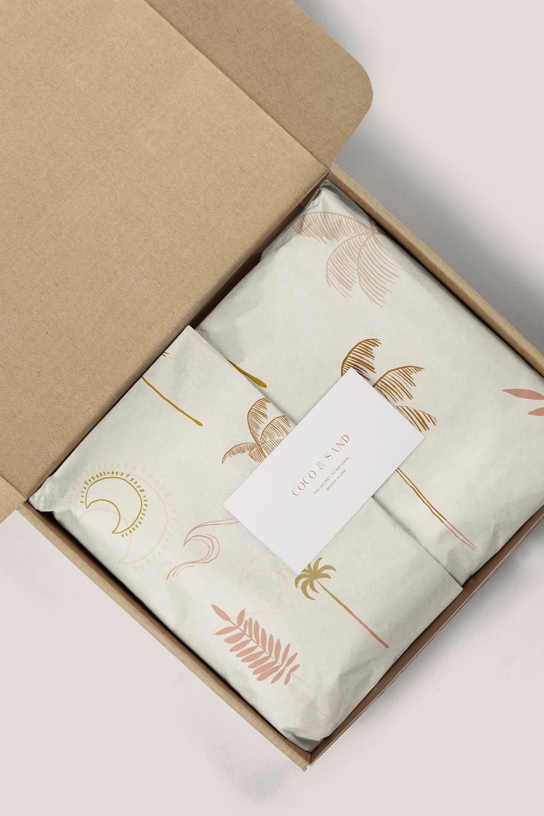 May include: A cardboard box with a white tissue paper lining. The tissue paper has a pattern of palm trees, leaves, and moons. A white business card with the text "Coco & Sand The Sweetest & Natural Beauty Balm" is on top of the tissue paper.