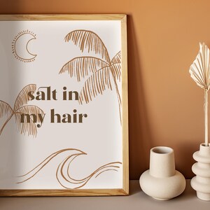 May include: A framed print with a white background and brown line drawings of palm trees, waves, and a crescent moon. The text "salt in my hair" is written in brown letters.