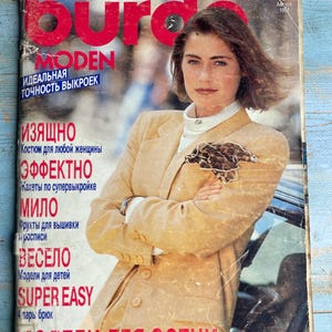 Op de afbeelding: Een vintage magazine cover met een vrouw in een beige blazer. De cover bevat Russische tekst, waaronder "MODEN" en "MODELLEN VOOR DE HERFST". Het tijdschrift is gedateerd augustus 1991.