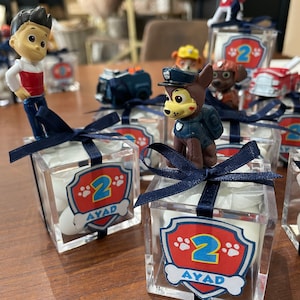 Peut inclure: Boîtes à dragées en plastique transparent avec un ruban bleu et un personnage de Pat'Patrouille sur le dessus. Les boîtes sont remplies de bonbons blancs et portent un logo Pat'Patrouille rouge et blanc avec le numéro 2 et le nom Ayad.