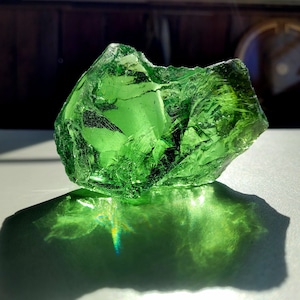 May include: A large, green, glass shard with a rough, irregular shape. The shard is reflecting light, creating a rainbow effect.