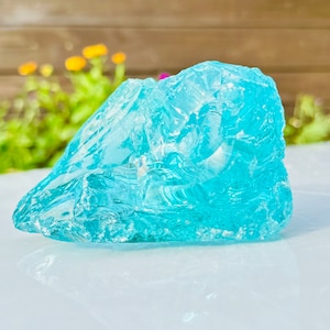 Op de afbeelding: Een groot, onregelmatig gevormd stuk turquoise glas. Het glas heeft een ruw, getextureerd oppervlak en lijkt doorschijnend, met licht dat erdoorheen schijnt. Het object staat tegen een vage achtergrond van groen en een houten hek.