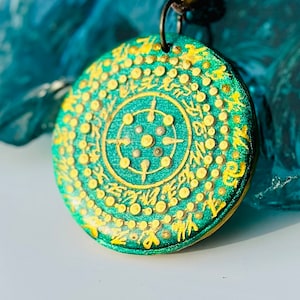 Puede incluir: Un colgante redondo verde con letras doradas y un diseño circular en el centro. El colgante está en un cordón de cuero marrón.