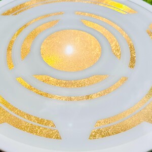 May include: A white decorative plate with a gold geometric design. The plate features concentric circles and angular shapes in gold leaf against a white background. The design is centered, creating a focal point. The plate is round.