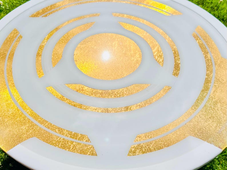 May include: A white decorative plate with a gold geometric design. The plate features concentric circles and angular shapes in gold leaf against a white background. The design is centered, creating a focal point. The plate is round.