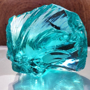 Op de afbeelding: Een groot, onregelmatig gevormd stuk doorschijnend, turquoise gekleurd glas. Het glas heeft een ruw, getextureerd oppervlak met wervelende patronen en interne breuken, die het licht vangen en reflecteren. Het algehele effect is een opvallende weergave van kleur en vorm.