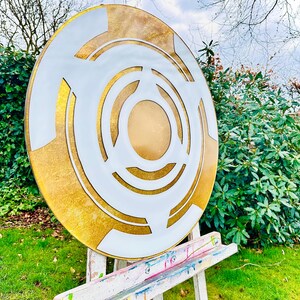 May include: A large, round, abstract art piece with a gold and white design. The artwork features concentric circles and geometric shapes. It is displayed on a white easel in a garden setting with green grass and foliage.