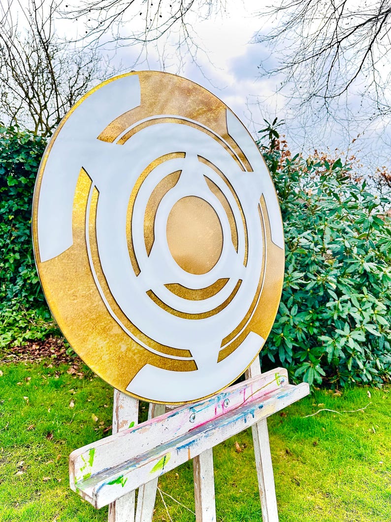May include: A large, round, abstract art piece with a gold and white design. The artwork features concentric circles and geometric shapes. It is displayed on a white easel in a garden setting with green grass and foliage.
