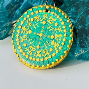 Puede incluir: Un colgante redondo, verde y dorado con un diseño circular y letras doradas. El colgante está en un cordón de cuero marrón.