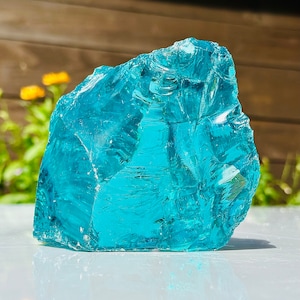 Puede incluir: Un gran trozo de vidrio azul transparente de forma irregular. El vidrio tiene una superficie rugosa y texturizada con muchas facetas, que reflejan la luz. El fondo es una vista borrosa de una valla de madera y vegetación.