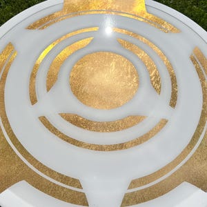 May include: A white and gold decorative plate with a circular design. The plate features a white outer rim and a gold center, with gold accents and a geometric pattern. The plate is likely made of ceramic or glass.