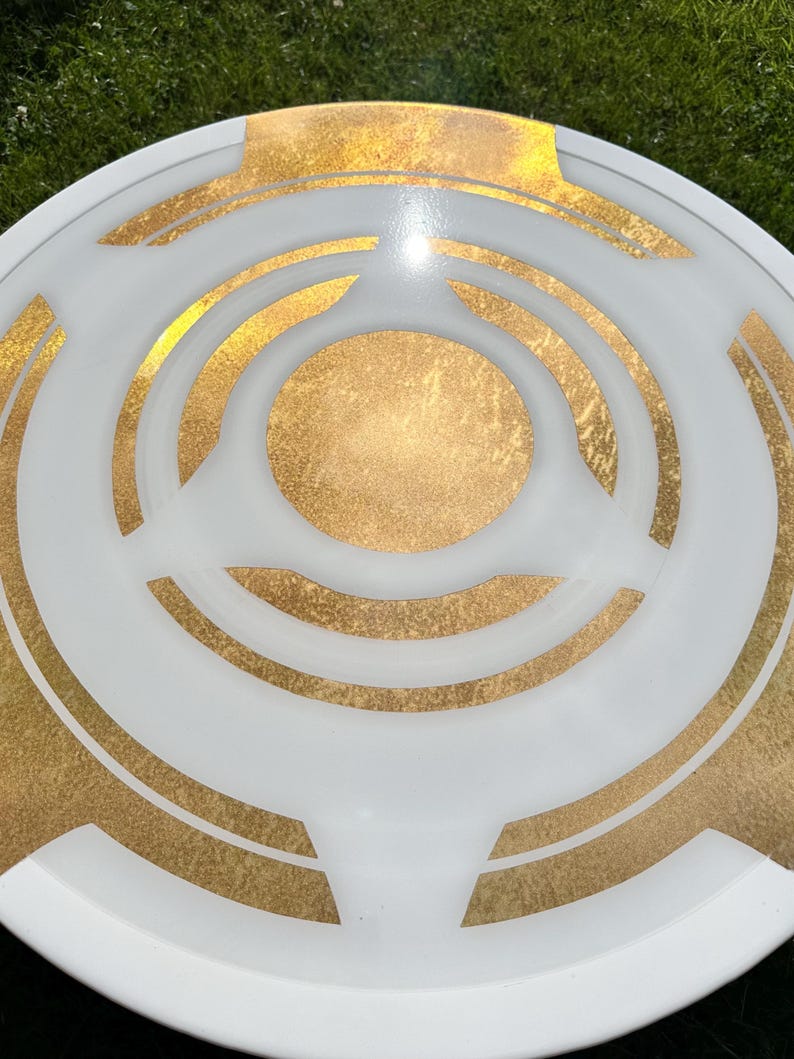 May include: A white and gold decorative plate with a circular design. The plate features a white outer rim and a gold center, with gold accents and a geometric pattern. The plate is likely made of ceramic or glass.
