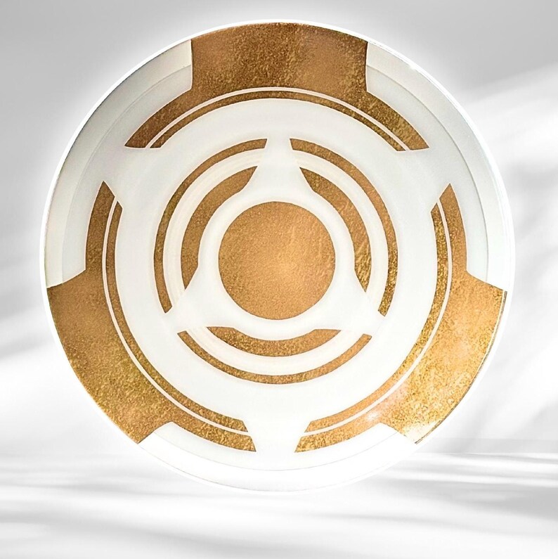 May include: A decorative white and gold plate with a circular design. The plate features a gold center and gold accents, with white geometric shapes. The plate is likely used for serving or display.