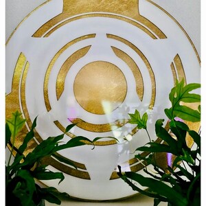May include: A decorative white plate with a gold geometric design. The plate features concentric circles and angular shapes in gold against a white background. Green plants frame the plate.