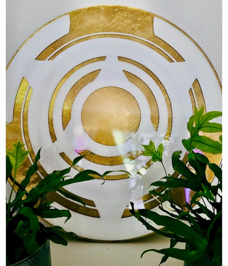 May include: A decorative white plate with a gold geometric design. The plate features concentric circles and angular shapes in gold against a white background. Green plants frame the plate.