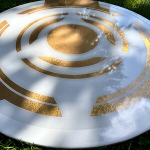 May include: A white and gold decorative shield with a circular design. The shield has a white base with gold geometric patterns and a gold center. The shield is round and appears to be made of a durable material.