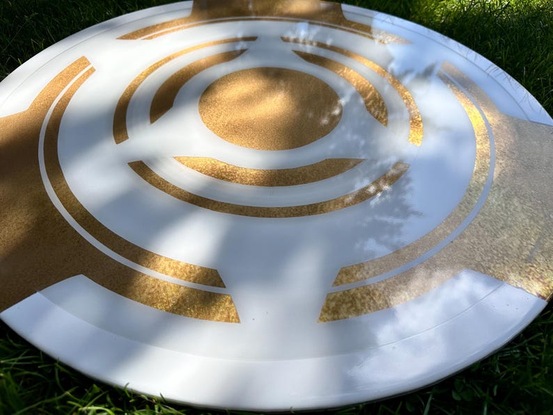 May include: A white and gold decorative shield with a circular design. The shield has a white base with gold geometric patterns and a gold center. The shield is round and appears to be made of a durable material.