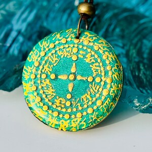 May include: A green and gold pendant necklace with a circular design featuring a central cross and a repeating pattern of dots and lines. The pendant is made of resin and has a glossy finish.