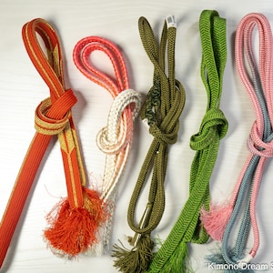 May include: A collection of five colorful obi jime, or kimono belts, displayed against a white background. The belts vary in color, including red with gold accents, pink and white, olive green, bright green, and pink and blue. Each belt is knotted.