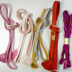 May include: An assortment of colorful Japanese obi cords, including pink, white, gold, silver, and red. Each cord is knotted and features decorative tassels. The cords are displayed on a white wooden surface.