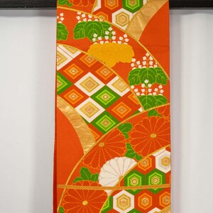 May include: An orange kimono fabric with a repeating pattern of stylized flowers, geometric shapes, and gold accents. The design includes orange, green, white, and yellow elements. The fabric is hanging from a black rod.