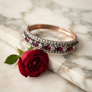 May include: A hinged bangle bracelet featuring a row of round-cut red rubies alternating with clear white stones, bordered by two rows of smaller clear stones. The bracelet is crafted from a metallic material and rests beside a single red rose.