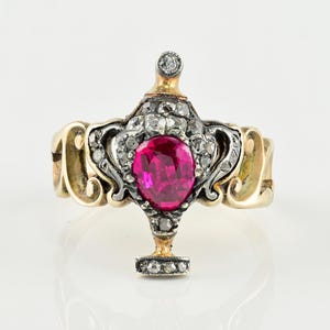 May include: An antique gold ring with a large, oval-cut ruby center stone. The ruby is surrounded by a halo of small diamonds. The ring has intricate detailing and a unique design, with a small diamond at the top.
