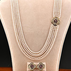 May include: A multi-strand pearl necklace with a decorative clasp and matching earrings. The clasp and earrings feature a gold-colored setting with a red floral design and small accent stones. The necklace and earrings are displayed on a beige jewelry stand.