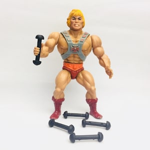 May include: A vintage He-Man action figure with a blonde wig, a gray and red chest armor, and a red loincloth. He is holding a black dumbbell in his right hand. There are three black dumbbells on the white surface below him.