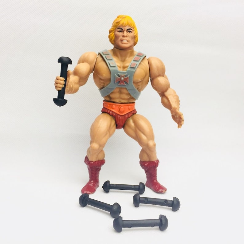 May include: A vintage He-Man action figure with a blonde wig, a gray and red chest armor, and a red loincloth. He is holding a black dumbbell in his right hand. There are three black dumbbells on the white surface below him.