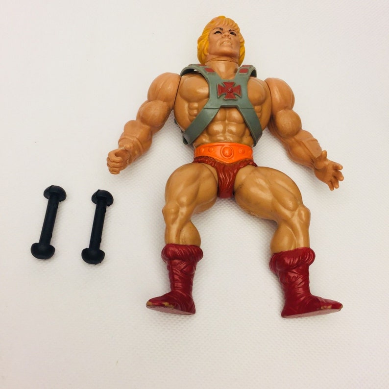 May include: A vintage He-Man action figure with a gray and orange chest armor, a red loincloth, and red boots. The figure has a blond hair and a muscular build. The figure is lying on its back with its arms outstretched.