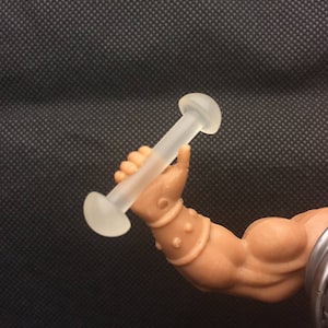 May include: A clear plastic dumbbell with rounded ends. The dumbbell is being held by a muscular arm.