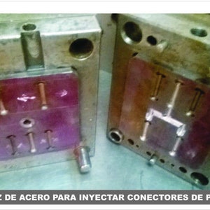 May include: Close-up of a metal mold with a red plastic insert. The mold is used to create plastic connectors for toy figures. The text "MATRIZ DE ACERO PARA INYECTAR CONECTORES DE PIERNAS MOTU" is visible on the bottom of the image.
