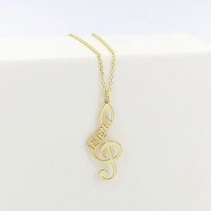 May include: Gold necklace with a treble clef pendant featuring the name "Elena" in cursive script.