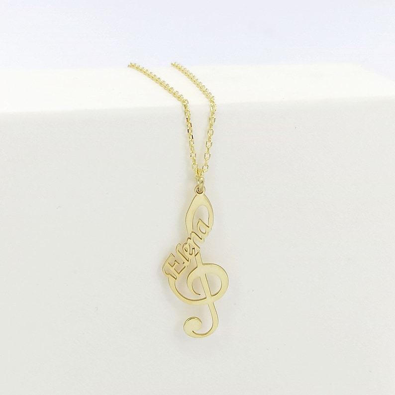 May include: Gold necklace with a treble clef pendant featuring the name "Elena" in cursive script.