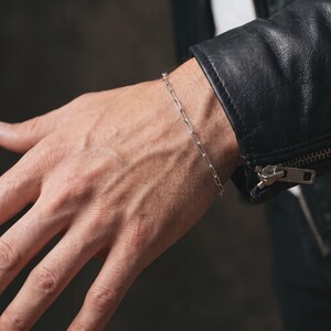 May include: A silver paperclip chain bracelet featuring elongated rectangular links. The bracelet is worn on the wrist, positioned near the cuff of a black leather jacket with a visible metal zipper pull and a silver-toned metallic finish.