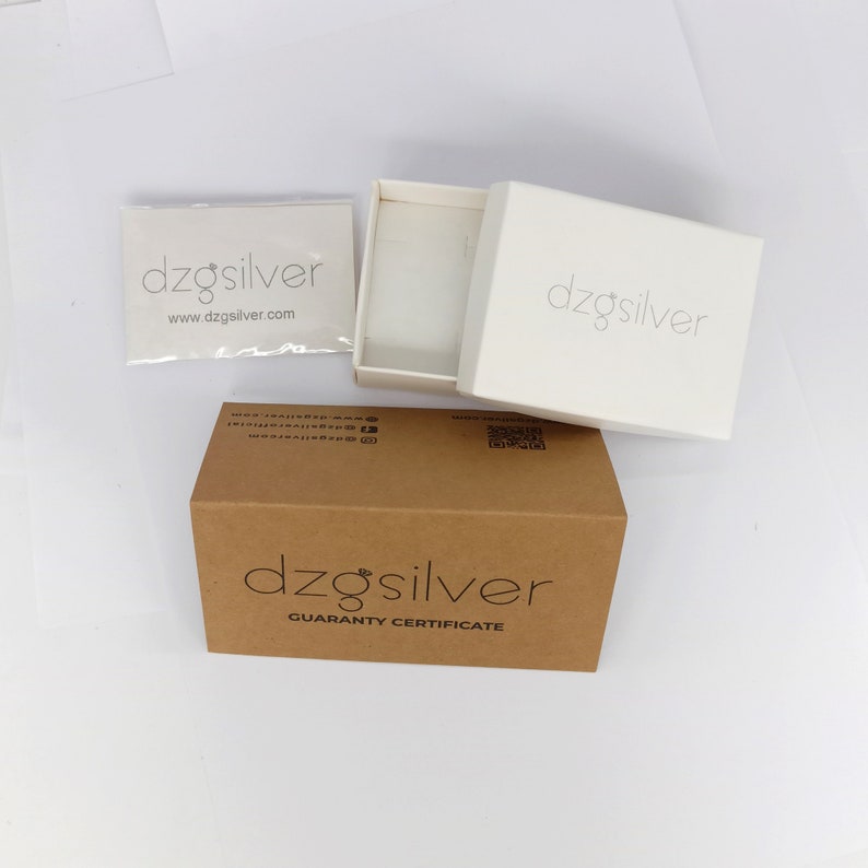以下が含まれることがあります： 茶色の段ボール箱に「dzgsilver」と「Guaranty Certificate」の文字が印刷されています。箱が開いており、中には「dzgsilver」の文字が書かれた白い箱が入っています。箱の上に「dzgsilver」と「www.dzgsilver.com」の文字が書かれた白いカードが置かれています。