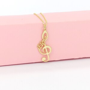 May include: Gold necklace with a treble clef pendant featuring the name "Elena" in cursive script.
