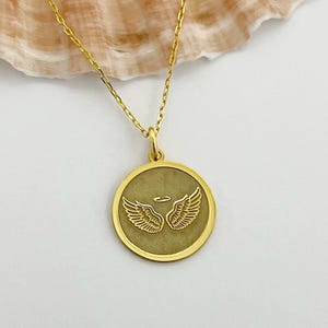 以下が含まれることがあります： 天使の羽根とハローが刻まれた丸いペンダントが付いたゴールドネックレス。