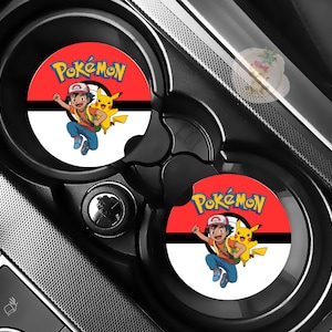 May include: Two black car cup holders with red and white circular coasters. Each coaster features the Pokémon logo and a cartoon image of Ash Ketchum and Pikachu. The coasters are designed to fit inside the car's cup holders.