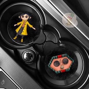 Pu&ograve; includere: Due sottobicchieri neri per auto con illustrazioni del film "Coraline". Uno mostra un personaggio con un cappotto giallo, l'altro un design a bottoni. I sottobicchieri sono posizionati nei portabicchieri di un'auto.