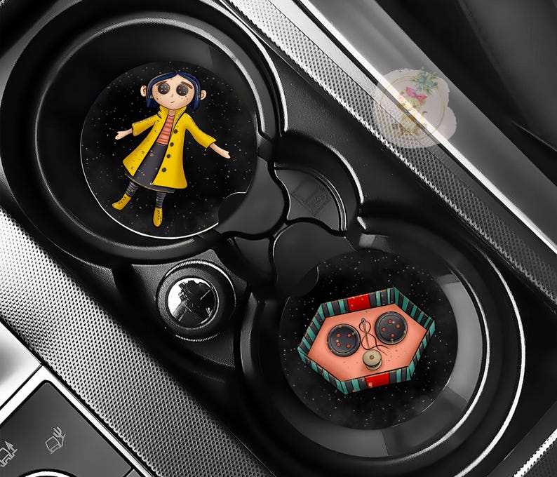 Pu&ograve; includere: Due sottobicchieri neri per auto con illustrazioni del film "Coraline". Uno mostra un personaggio con un cappotto giallo, l'altro un design a bottoni. I sottobicchieri sono posizionati nei portabicchieri di un'auto.