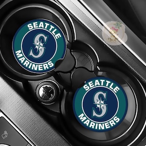 May include: Two car coasters with the Seattle Mariners logo, featuring a teal and navy blue color scheme. The coasters are circular and fit into a car's cup holders. The logo includes the team name and a stylized letter "S".