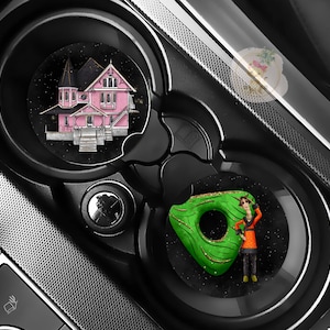 Pu&ograve; includere: Portabicchieri per auto neri con due sottobicchieri circolari. Uno presenta un'illustrazione di una casa rosa, l'altro una ciambella verde con una persona. I sottobicchieri sono su uno sfondo nero con effetto stellato.