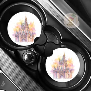 May include: Two white car coasters featuring a watercolor illustration of a fairytale castle with pink, yellow, and blue hues. The coasters are set inside a black car console cup holder.