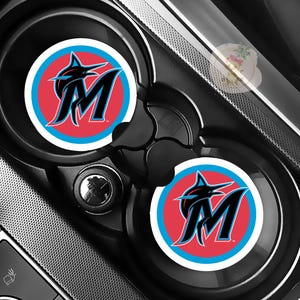 May include: Two black car cup holder coasters featuring the Miami Marlins logo. The logo includes a stylized marlin, the letter "M", and a red, blue, and white color scheme. The coasters are set in a car's cup holders.
