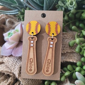 Puede incluir: Un par de pendientes colgantes con una pelota de s&oacute;ftbol amarilla con costuras rojas unida a un bate de b&eacute;isbol de madera clara. El bate tiene grabado el nombre Chloe y presenta dos peque&ntilde;os detalles rectangulares blancos en el mango.